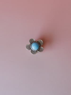 A silver flower-shaped ring with a rounded turquoise stone center sits centered against a muted dusty rose background — the kind of piece that looks like it came from a vintage market and somehow goes with everything.