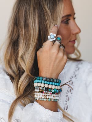 A close-up of stacked beaded bracelets and layered statement rings on a model's hand — turquoise stones, cream wood beads, and silver flower rings together in that effortless 'collected over time' way.