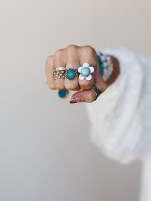 A fist of rings — stacked silver, gold daisy, turquoise stone flower, and a chunky white enamel bloom — worn together the way someone who actually loves jewelry does it, no rules, just instinct.