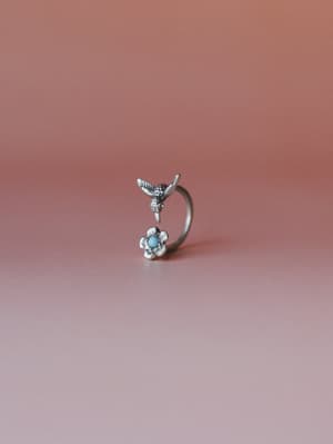 A delicate silver open-band ring with a hummingbird perched above a flower centerpiece set with a small turquoise stone, shot solo against a warm dusty rose backdrop — the kind of piece you reach for every single day without thinking.