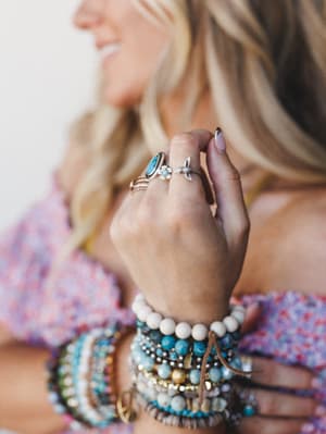 A close-up lifestyle shot of a model's hand adorned with stacked silver rings — including a turquoise stone ring and a delicate floral-detail ring — layered over a wrist piled high with beaded bracelets in turquoise, cream, and gold tones, all agains