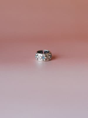 A wide-band silver ring with embossed daisy florals and turquoise stone centers sits centered against a warm dusty rose gradient background — the kind of piece that looks like it came from a vintage market but feels totally intentional.