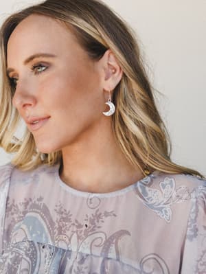 A close-up lifestyle shot highlighting silver crescent moon drop earrings against soft, effortless styling — the model's relaxed gaze and dreamy paisley top give the whole thing a quietly romantic, celestial feel.