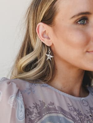 A close-up lifestyle shot spotlighting a silver star drop earring worn by a blonde model, styled with a soft lavender paisley top — the kind of detail shot that makes you pause mid-scroll.