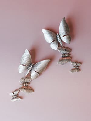 A pair of cascading silver butterfly drop earrings laid against a dusty rose background — each earring features a large statement butterfly at the top with two progressively smaller ones trailing below, all with beautifully textured, wing-etched deta
