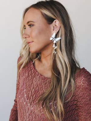 A close-up portrait shot showcasing cascading silver butterfly drop earrings styled against a dusty rose open-knit sweater — soft, dreamy, and the kind of detail that makes the whole outfit.