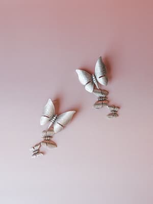 A pair of silver butterfly drop earrings laid flat against a dusty rose background — each earring features a cascade of three butterflies in descending sizes, with a brushed metallic finish that catches light softly.