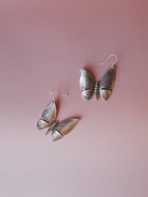 A pair of large silver butterfly drop earrings laid against a dusty rose background — textured, worn-in metal with beautifully detailed wings that feel like something you'd find at a vintage market and never take off.