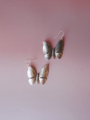A pair of oversized silver butterfly drop earrings laid against a dusty rose background — textured, hand-finished wings with a slightly antiqued quality that feels collected, not costume.