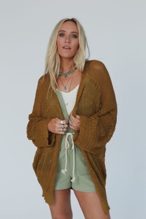 A woman with long blonde hair wears the Brenden Knit Netted Cardigan in brown over a white top and green shorts, accessorized with layered necklaces and rings, standing against a plain light background.