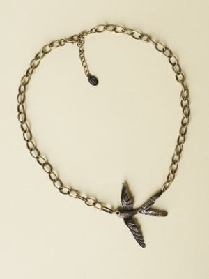 A bronze-toned chain necklace with a detailed flying bird pendant — the kind of piece that feels like it has a story, laid flat against a soft cream background.