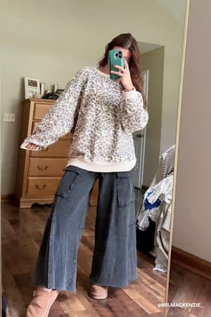 A woman stands in front of a mirror taking a selfie, wearing the Game On Cargo Wide Leg Pant in black paired with a loose leopard-print sweater and tan shoes. The room has wooden floors, a dresser, and clothes visible in the background.