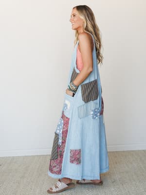 A sleeveless denim maxi jumpsuit with wide-leg silhouette covered in mixed patchwork panels — paisley, stripe, and daisy prints — styled over a coral tank with stacked beaded bracelets and white buckle sandals, shot in profile against a clean white s