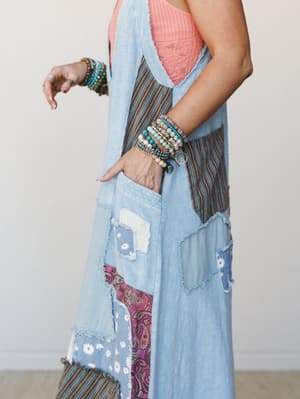 A side-profile close-up of a patchwork denim maxi vest layered over a coral ribbed tank, styled with an armful of stacked beaded bracelets — the kind of outfit that looks like it came together effortlessly but very much did not.