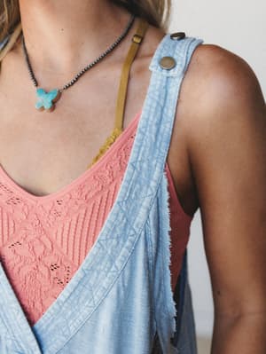 A close-up chest-and-shoulder shot capturing the layered boho texture of a washed denim overall strap over a dusty rose lace tank, finished with a turquoise butterfly pendant necklace — the kind of outfit that looks effortless because it genuinely is