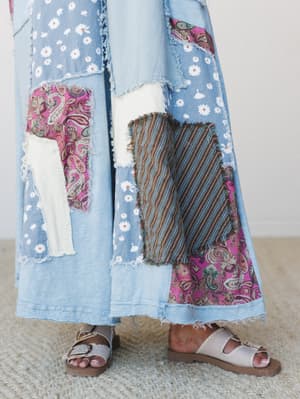 A close-up of wide-leg denim pants covered in mixed patchwork — daisy print, paisley, stripe, and raw-edge fabric panels — worn with buckle slide sandals on a woven rug, giving off serious vintage thrift-store-find energy in the best way.