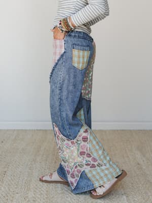 A side-profile shot of wide-leg patchwork denim jeans styled with a striped long-sleeve top and flat sandals — the kind of pants that do all the talking, covered in plaid, floral, and polka dot fabric patches with a standout star appliqué at the hem.