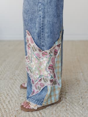 A close-up detail shot of wide-leg acid wash denim jeans featuring a star-shaped patchwork appliqué at the hem, layered with mixed fabric patches in floral, plaid, and abstract prints — the kind of jeans that make people stop you mid-errand to ask wh