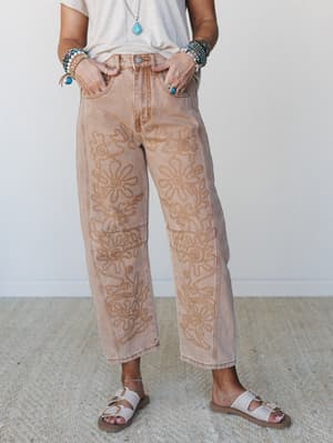 A mid-shot of wide-leg caramel-washed jeans covered in tonal floral embroidery, styled with a cream tee, stacked boho bracelets, a turquoise pendant necklace, and white buckle sandals — the kind of pants that do all the talking.