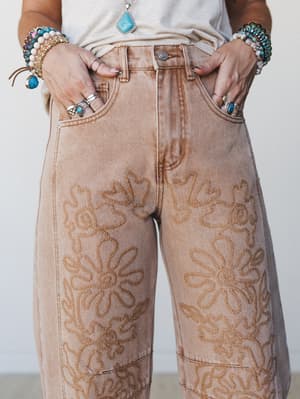 A close-up detail shot of wide-leg denim pants in a warm washed rust tone, covered in tone-on-tone floral embroidery — styled with stacked boho bracelets, silver rings, and a turquoise pendant that feel like they were collected, not coordinated.