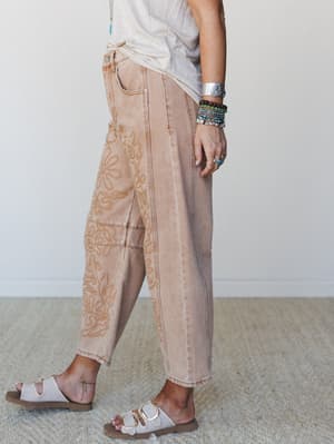 A side-angle shot of acid-washed rust denim barrel-leg pants with tonal floral embroidery, styled with a knotted cream tee, stacked bracelets, and white buckle sandals — the kind of outfit that looks effortless but is clearly very much on purpose.