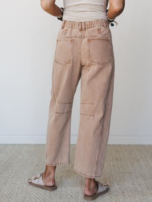 A back-view shot of relaxed wide-leg denim pants in a warm sandy rust wash, styled with a cream tank and flat sandals — the kind of pants that feel like a soft answer to everything you need to do today.