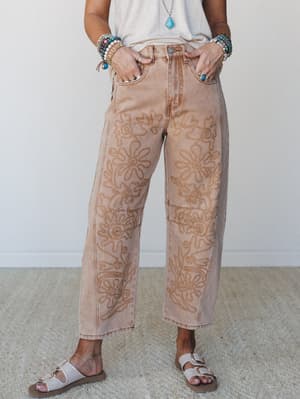 Acid-washed barrel-leg jeans in a warm sandy rust tone, covered in tonal floral embroidery, styled with a cream tee, stacked beaded bracelets, a turquoise pendant, and white buckle sandals — the kind of outfit that looks effortless but very much on p