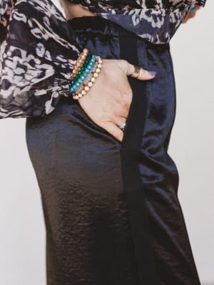 Person wearing a patterned top and dark pants with colorful bracelets on a plain background