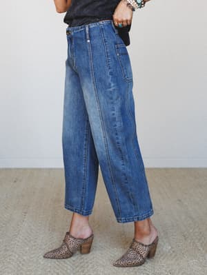 A cropped side-angle shot showing wide-leg medium wash denim jeans styled with leopard print mules and stacked boho jewelry — relaxed and lived-in with a cool vintage energy.
