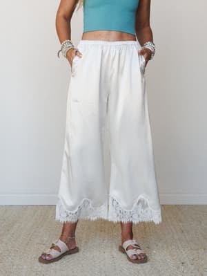 Wide-leg satin pants in soft ivory with a scalloped lace hem trim, styled with a cropped teal top, stacked bead bracelets, and strappy sandals — easy and a little dreamy.