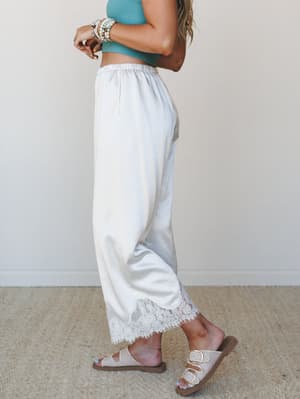Wide-leg satin-finish pants in champagne with a delicate lace hem finish, styled with a teal ribbed crop top, stacked bead bracelets, and double-buckle sandals — effortless and a little romantic, like a beach vacation that turned into a lifestyle.
