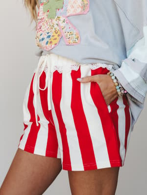 A close crop of red and cream wide-stripe shorts with a lace-trim waistband and drawstring tie, styled with a patchwork butterfly graphic sweatshirt and beaded bracelet — playful, summery, and a little nostalgic.