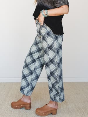 A side-profile shot of wide-leg plaid pants in sage and navy, styled with a black textured knit top, turquoise stacked bracelets, and warm brown clogs — the kind of outfit that looks effortless but was definitely thought about.