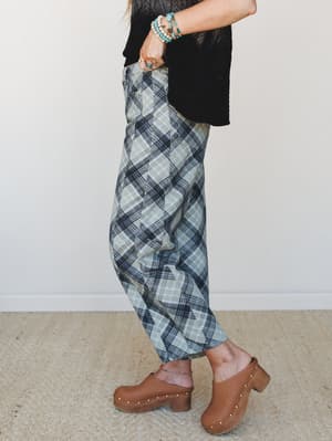 A side-profile shot of wide-leg plaid pants in sage and navy, styled with a black knit top and warm brown clogs — the kind of outfit that looks effortless but is clearly very thought out.