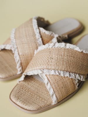 A pair of woven raffia slide sandals with crisscross straps and delicate crochet lace trim, resting casually on a soft sage-yellow surface — the kind of shoe that makes a sundress feel complete.