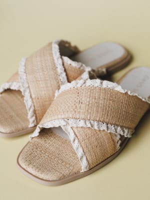 A pair of woven raffia slide sandals with crochet-trimmed crossover straps, shot close-up against a soft yellow-green backdrop — the kind of sandal that makes every summer outfit feel a little more intentional.