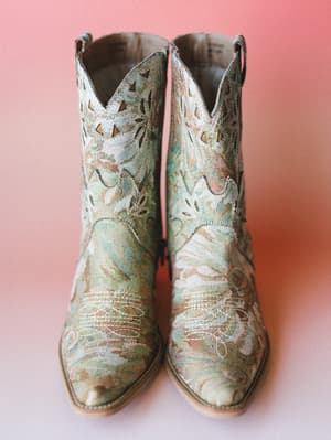 A pair of floral brocade cowboy booties in sage and cream sit centered against a warm coral-pink background — the kind of boots that make the whole outfit.