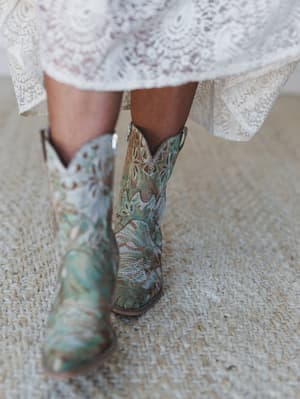 A close-up shot of intricate embroidered cowboy boots in sage and turquoise paired with a flowy lace dress hem — the kind of combination that feels equally at home at a barn wedding or a weekend festival.