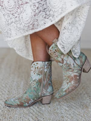 A close-up of floral-embroidered western booties in sage and dusty rose, peeking out from beneath the hem of a lace midi dress — the kind of styling moment that feels both intentional and completely effortless.