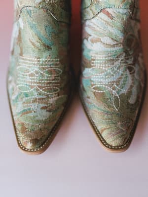 A close-up overhead shot of a pair of green brocade cowboy booties, their floral jacquard fabric and white topstitching detail doing all the talking against a soft blush surface.