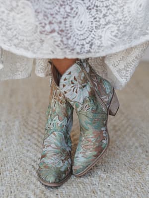 A close-up detail shot of floral brocade cowboy booties in sage and dusty teal, peeking out beneath the hem of a white lace dress — the kind of boot that makes the whole outfit.