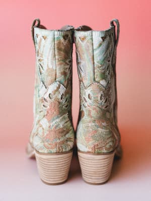 A pair of tapestry-fabric cowboy booties shot from the back against a warm coral backdrop — the floral brocade, stacked heel, and western stitching details make them feel like something you'd find at a vintage market and never put down.