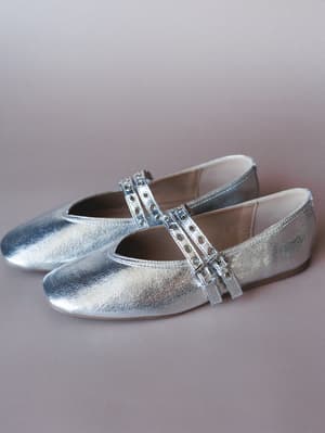 A pair of silver metallic Mary Jane flats shot against a soft mauve backdrop — the double eyelet straps give them just enough edge to feel intentional rather than precious.
