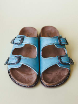 A pair of sky-teal double-strap leather slides shot from above against a soft cream backdrop — the kind of sandals that go from the farmers market to the back patio without a second thought.