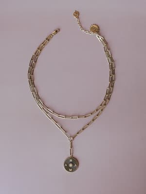 A layered gold paperclip chain necklace with an engraved 'MAMA' medallion pendant, laid flat on a dusty mauve background — the kind of piece that feels personal the moment you put it on.