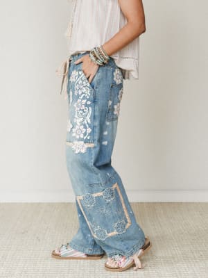 A side-profile shot of wide-leg denim jeans covered in floral appliqués, embroidery, and lace patchwork panels — styled with a striped tie-front top, stacked bracelets, and beaded sandals for a lived-in boho look.