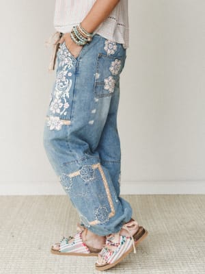 A side-profile shot of wide-leg denim jeans covered in floral appliqué, embroidery, and patchwork details — styled with a striped top, stacked bead bracelets, and whimsical embellished sandals for a fully lived-in boho look.