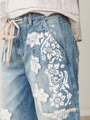 A close-up detail shot of light-wash denim jeans covered in white floral appliqué patches and painted floral scroll work, paired with a striped lace-trim top with a trailing tie — the kind of jeans that make people stop and ask where you got them.