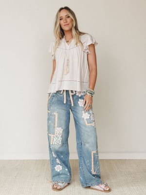 A breezy full-body shot of a model in wide-leg patchwork denim jeans and a flowy striped ruffle-sleeve top, styled with layered boho jewelry against a soft neutral studio wall — the kind of outfit that looks effortless but was definitely thought abou
