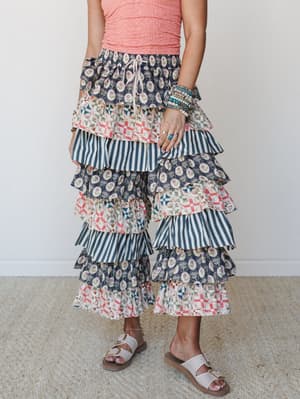 A tiered ruffle wide-leg pant in a mixed print patchwork of navy floral block print, stripe, and quilt-inspired geometric panels, styled with a coral textured top, stacked boho bracelets, and flat buckle sandals — the kind of statement bottom that do